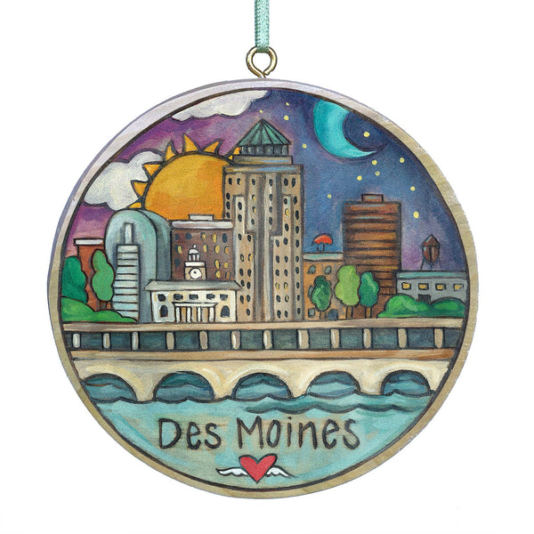 A circular ornament featuring a colorful illustration of the Des Moines skyline, including the sky with sun and moon, buildings, and the text 'Des Moines'.