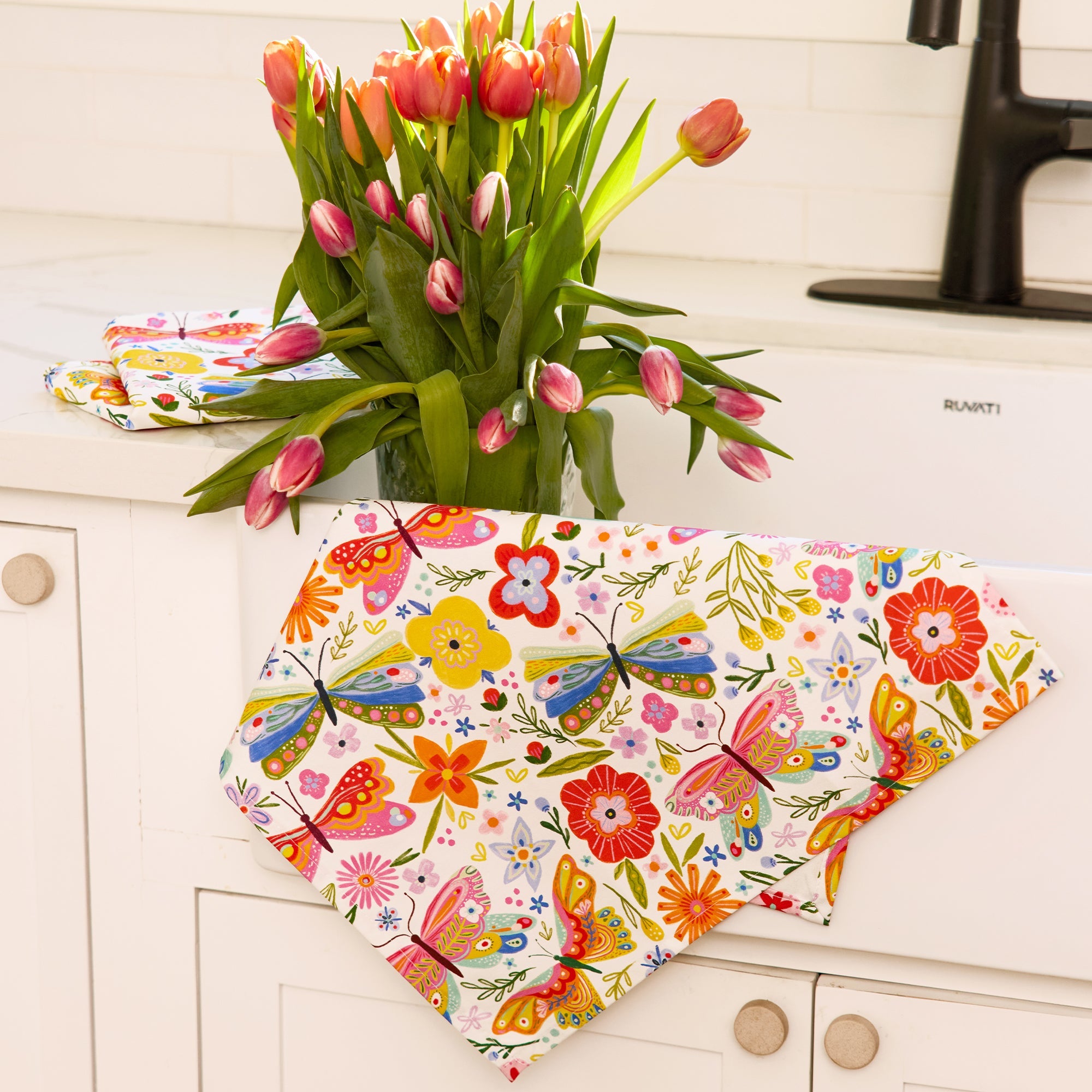 Colorful butterfly and floral patterned tablecloth on a white cabinet with tulips in the background.
