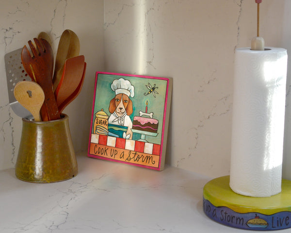 Kitchen counter with wooden utensils, a paper towel holder, and a dog chef wall plaqe.
