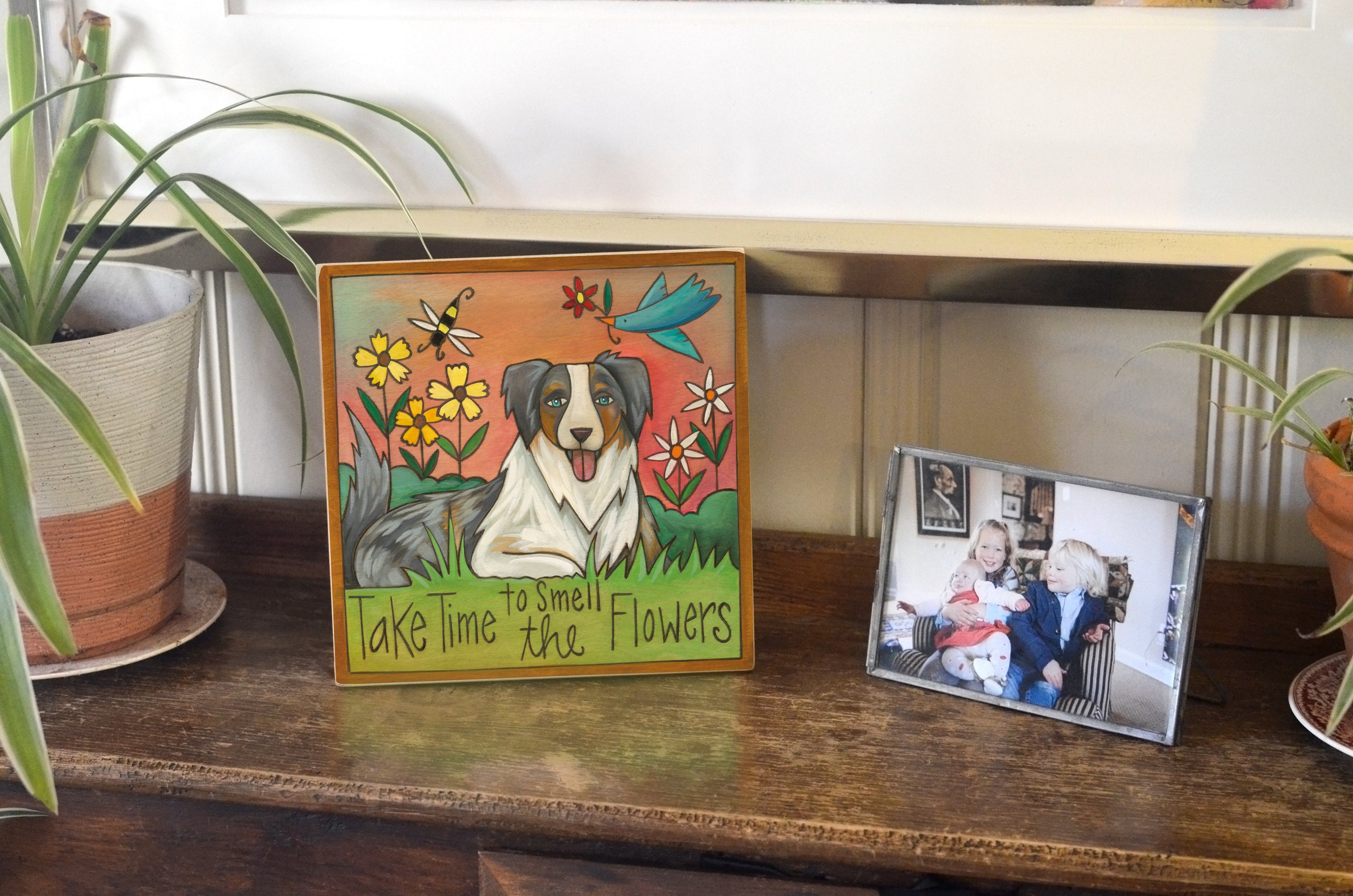 Decorative plaque with a dog illustration and text on a wooden surface with plants and a family photo.