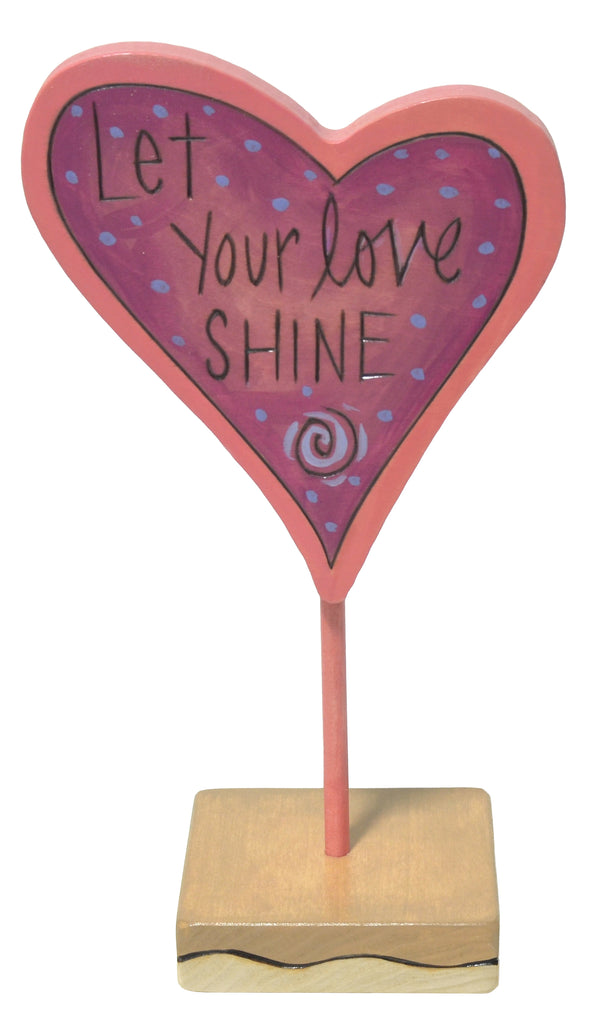 Heart-shaped sign with 'Let Your Love Shine' text on a stand