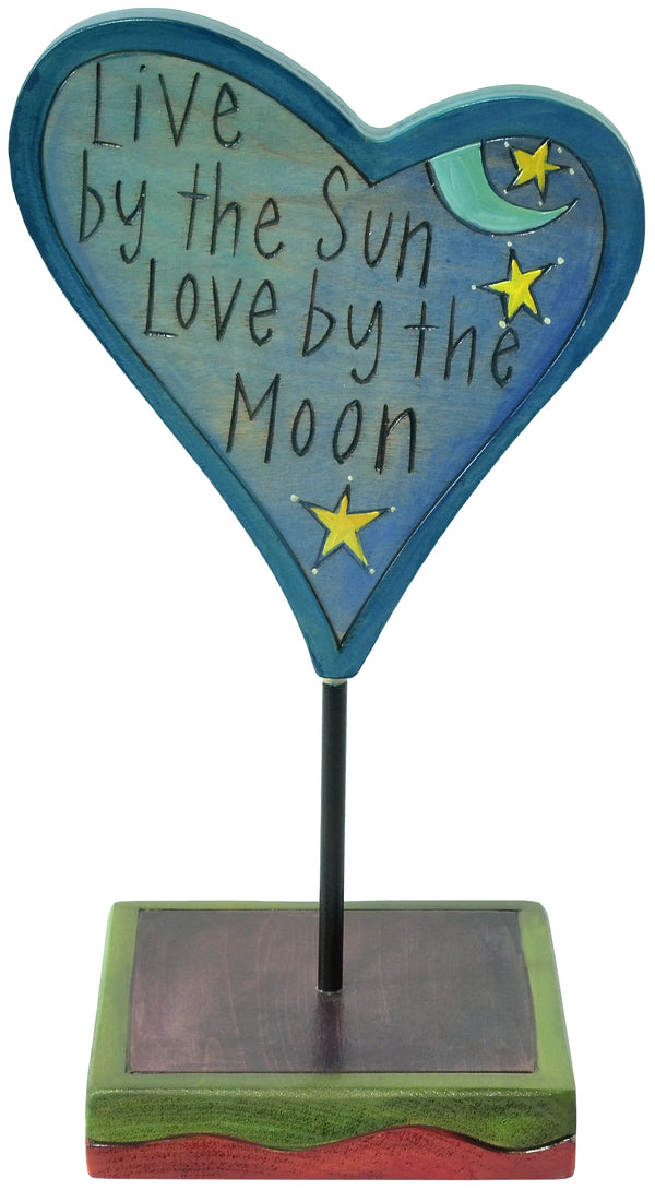 Heart-shaped blue sculpture with 'Live by the Sun, Love by the Moon' text on a stand.