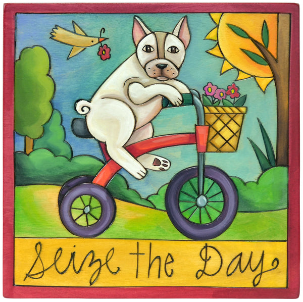 Colorful wood plaque of a white French Bulldog riding a tricycle with a basket, surrounded by nature, and the text 'Seize the Day'.