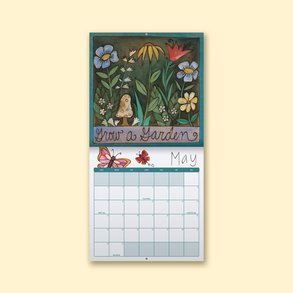A hanging monthly calendar page with a decorative floral illustration at the top and a May calendar grid below, shown against a pale cream background.