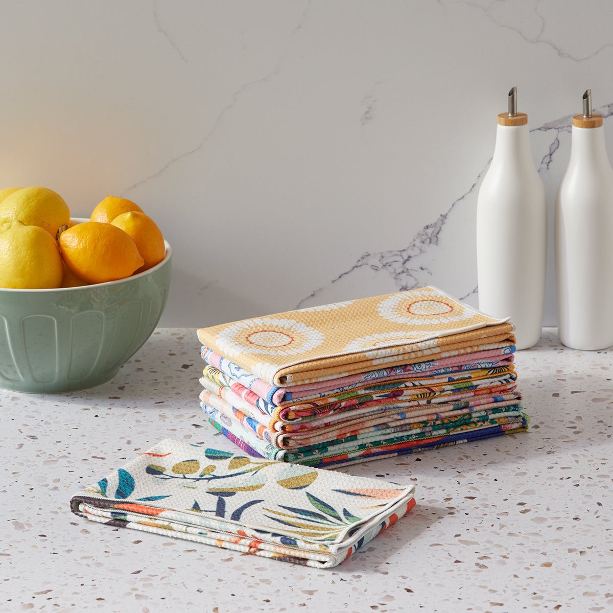 Stack of colorful folded towels on a kitchen counter with a bowl of fruit and bottles in the background.