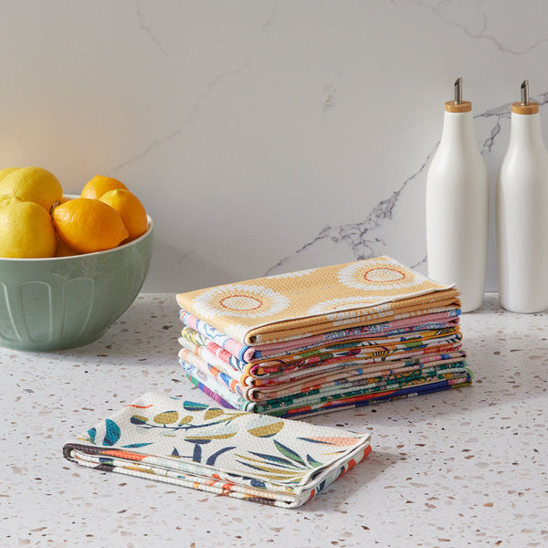 Stack of colorful folded towels on a kitchen counter with a bowl of fruit and bottles in the background.