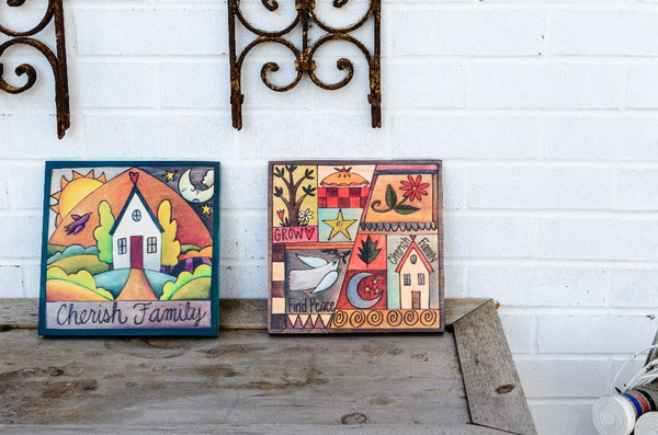 "Fly Home" Plaque – A vibrant scene of a house with a bird, moon, and sun in the sky above displayed against a white brick wall